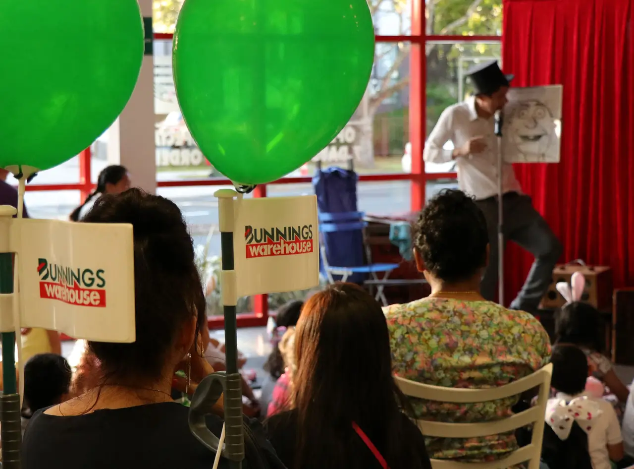 Crowd at Bunnings Australia relishes Andy Wonder's comedy magic amid green balloons, red curtain backdrop.