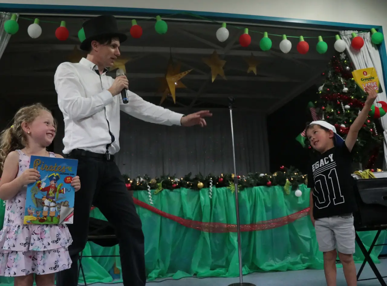 Magician engages with delighted kids amid festive stage setup.